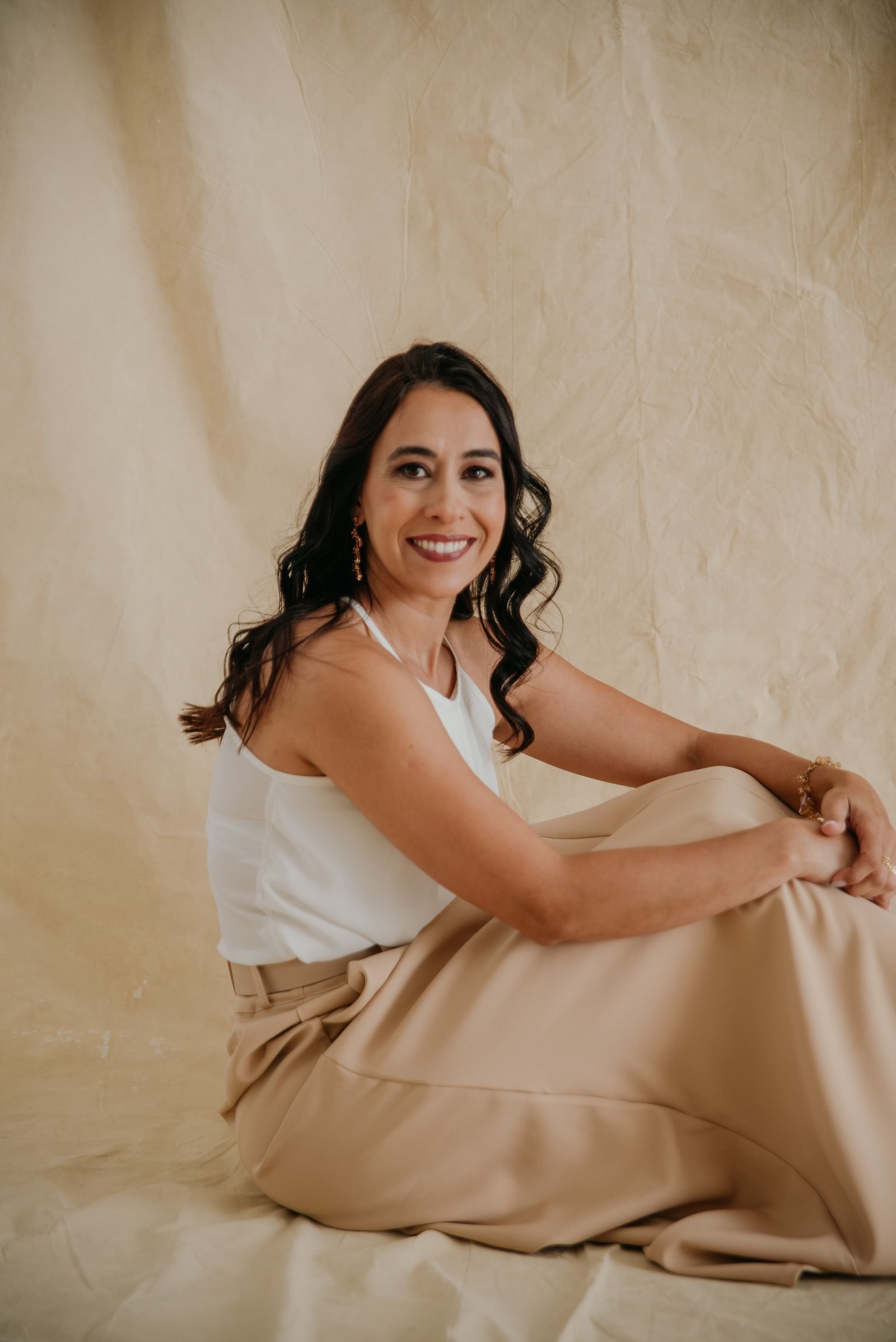 Fundadora da Florada de Ipê sorrindo, sentada sobre um tecido creme, vestindo blusa branca e saia bege